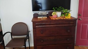 Una habitación con un televisor montado en la pared, un aparador de madera con libros y plantas, una silla marrón y una puerta roja.