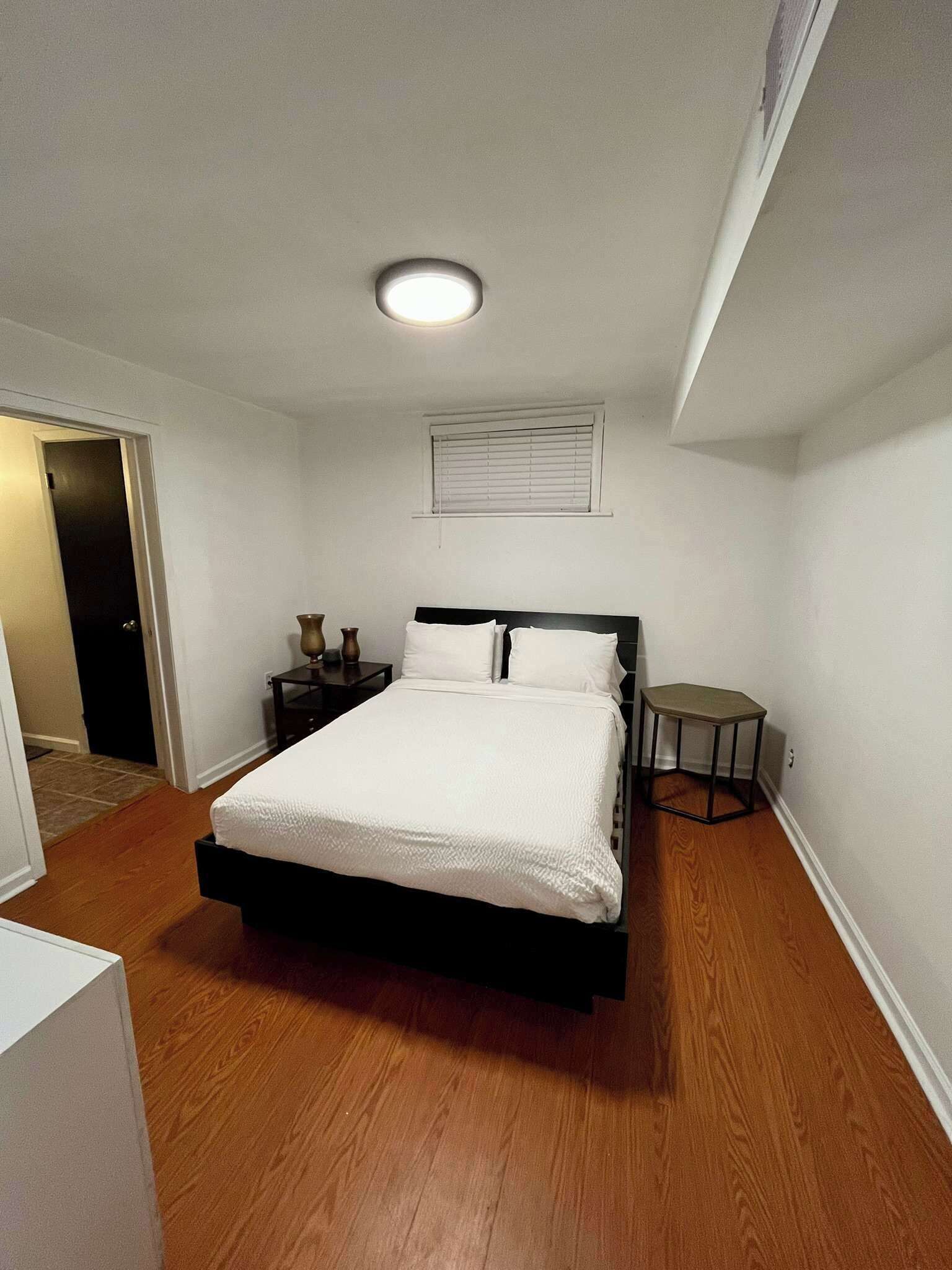 Un dormitorio con cama, mesitas de noche, una ventana con persianas y una lámpara en el techo.