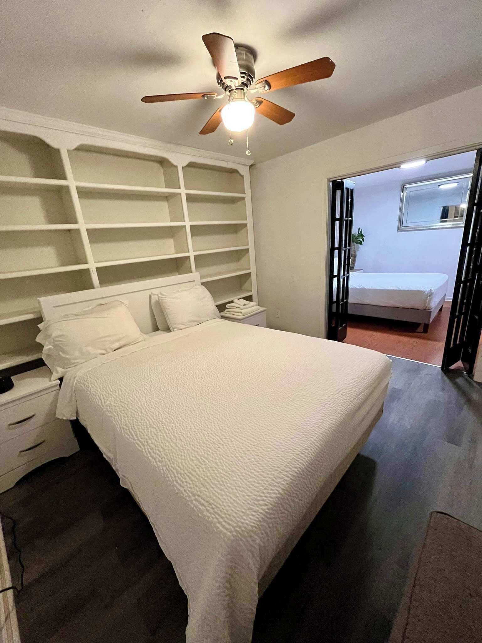 Une chambre à coucher comprenant un lit, un ventilateur de plafond, une table de chevet, une commode et un dressing avec des étagères.