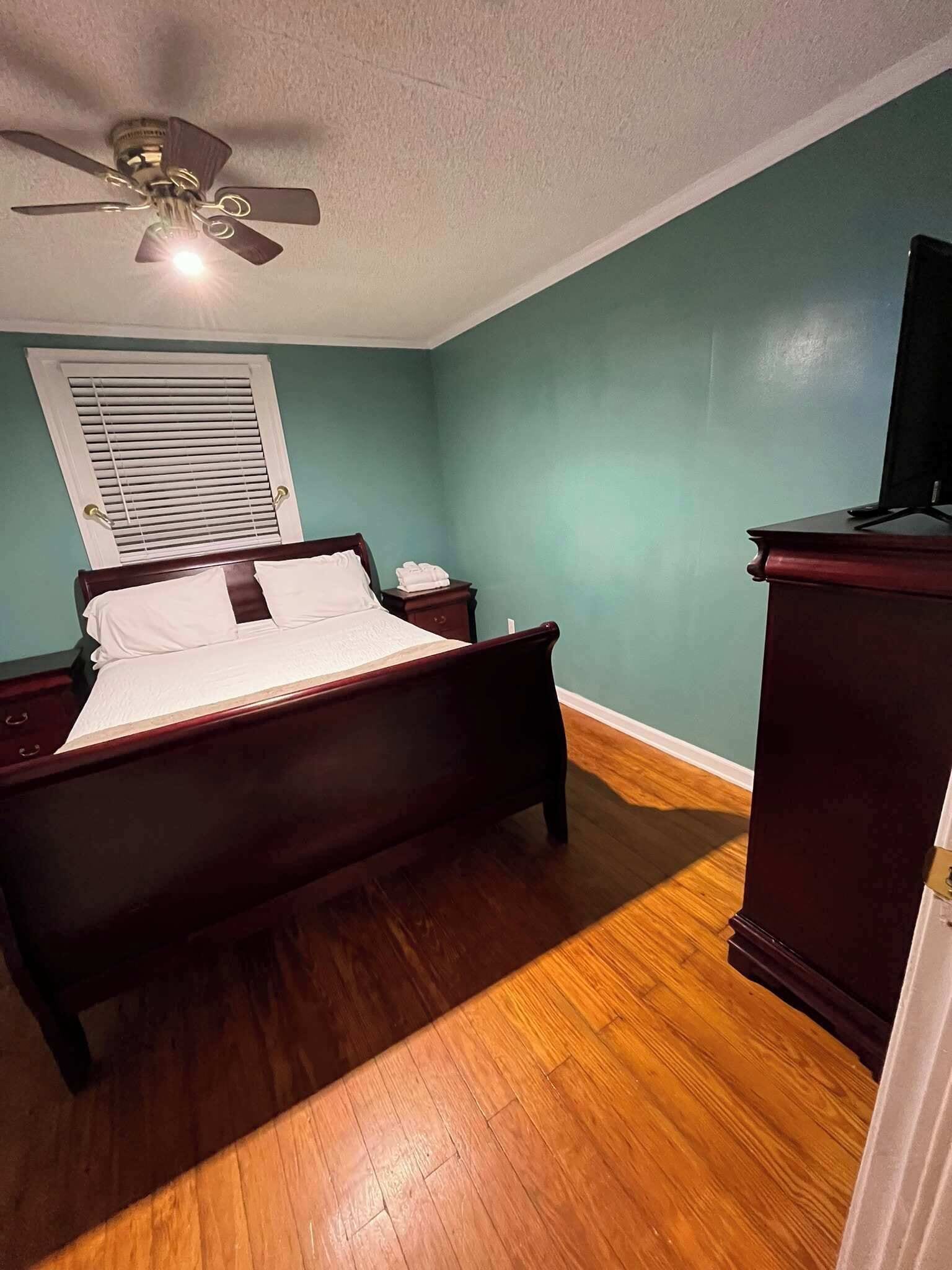 A bedroom with a wooden bed, a ceiling fan, a nightstand, a dresser, and a window with blinds.