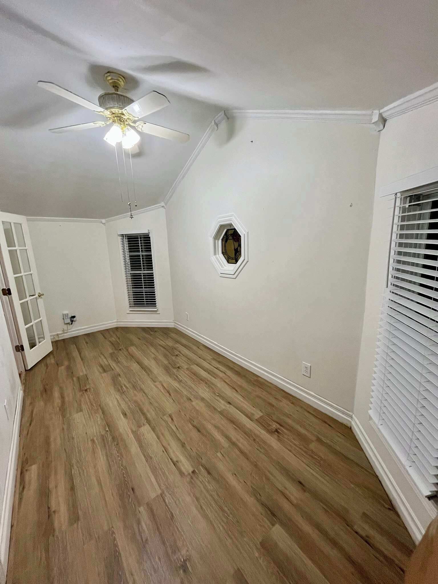 Ein Zimmer mit Holzboden, Deckenventilator, einem kleinen Fenster mit Zierrahmen und einer Tür mit Glasfeldern.