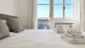 Une chambre d’hôtel avec un grand lit, des oreillers à carreaux, un radiateur et une vue sur la ville par la fenêtre.