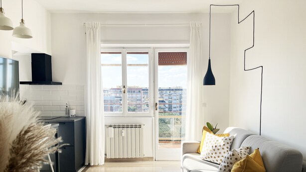 Un salon moderne avec un canapé, un fauteuil jaune et une table à manger. Il y a également un coin cuisine avec une cuisinière et des placards. La pièce est dotée de grandes fenêtres offrant une vue sur les bâtiments environnants.
