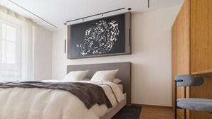 A modern bedroom with a large bed, a headboard, a grey chair, and a wall-mounted artwork. - Ma.maison (Guémar)