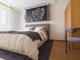 Un dormitorio moderno con una cama grande, un cabecero, una silla gris y una obra de arte colgada en la pared.