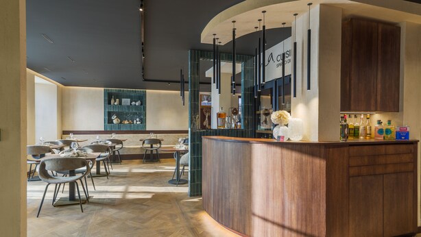 A modern bar area with a curved wooden counter, seating with tables and chairs, and a decorative wall with shelves. - Ma.maison (Guémar)