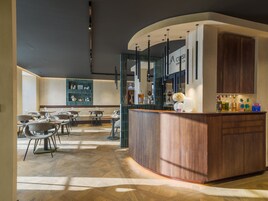 Zona de bar moderna con una barra de madera curvada, asientos con mesas y sillas, y una pared decorativa con estantes.