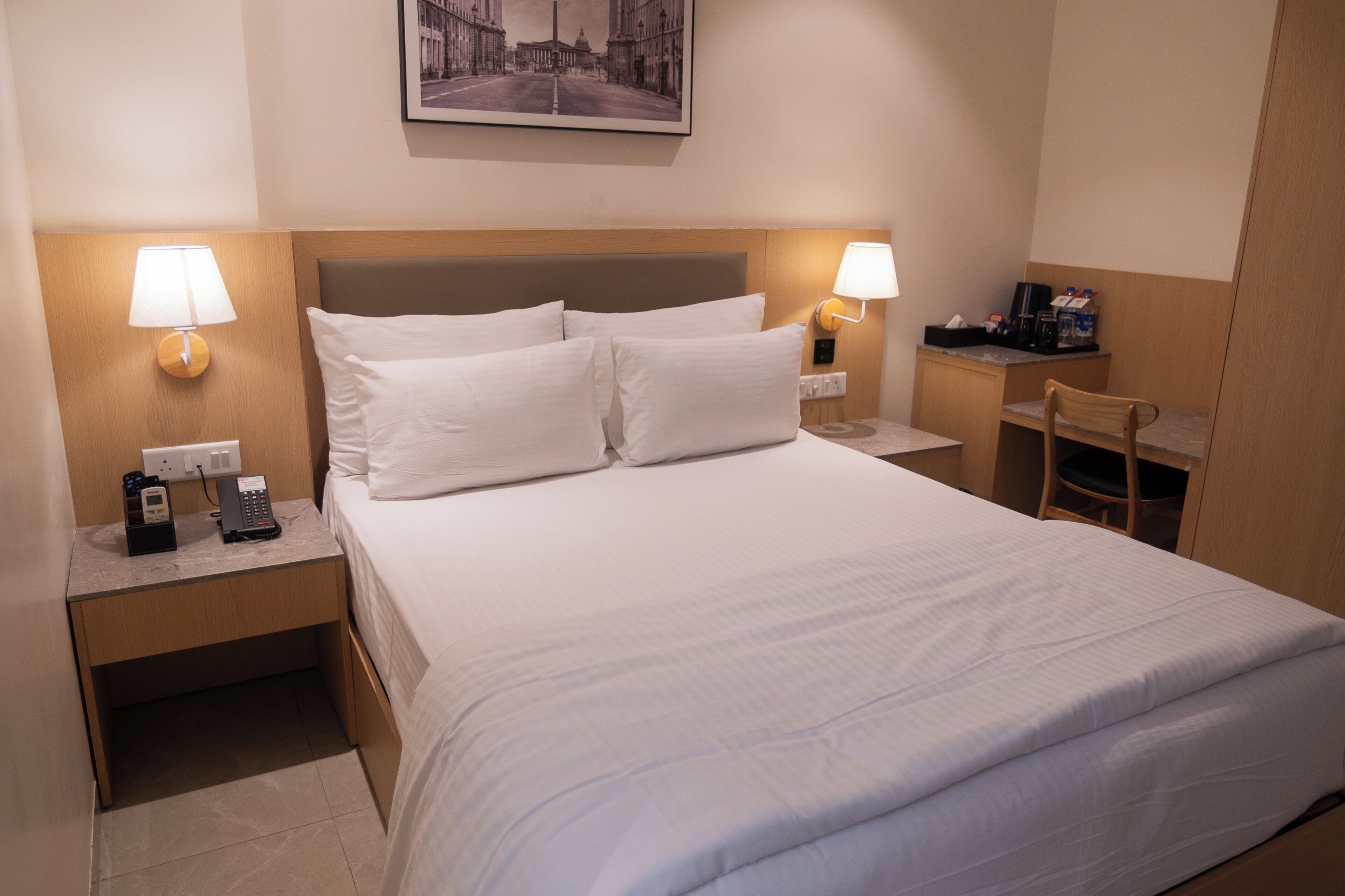 Habitación de hotel con cama, dos lámparas de noche, escritorio con silla, teléfono y un cuadro en la pared.
