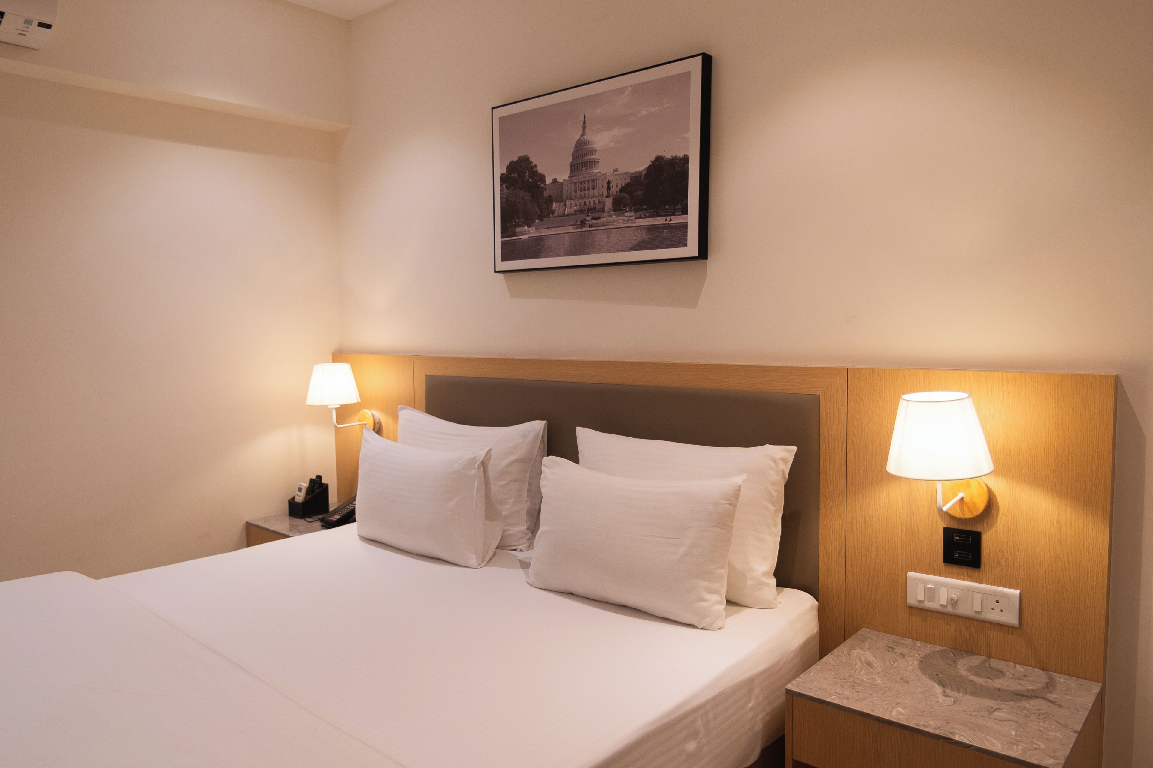 Una habitación de hotel con una cama, dos lámparas de noche, una mesita de noche, un enchufe en la pared y un cuadro con una vista de un edificio.