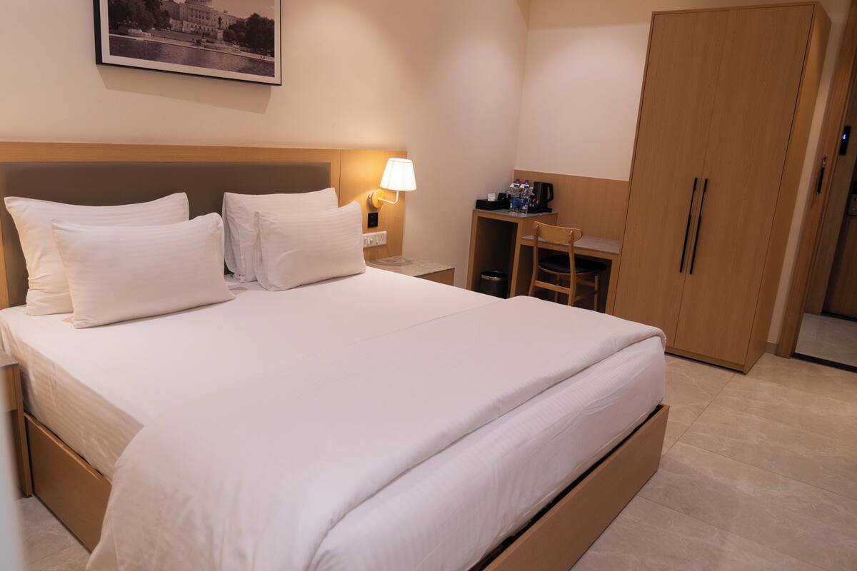 A hotel room with a bed, a nightstand with a lamp, a desk with a chair, and a wardrobe.