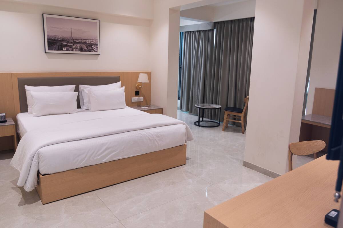 A hotel room with a bed, a nightstand, a chair, a small table, a lamp, a picture on the wall, and a view of the outside through a window.