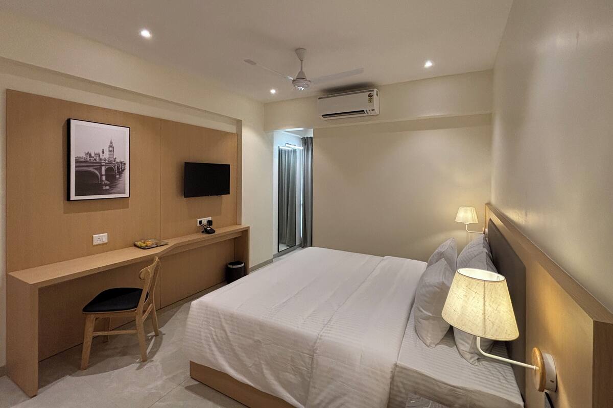 A hotel room with a bed, a desk, a chair, a TV, a lamp, and an air conditioning unit.