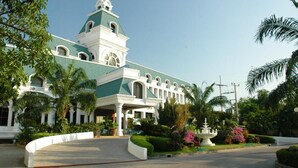 Un edificio grande e histórico, con techo verde y fachada blanca, rodeado de exuberante vegetación y palmeras.