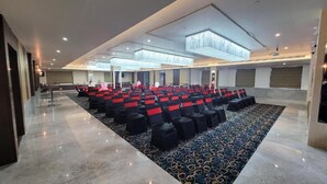 Uma sala de conferências com fileiras de cadeiras pretas com detalhes em vermelho, um carpete estampado e um lustre.