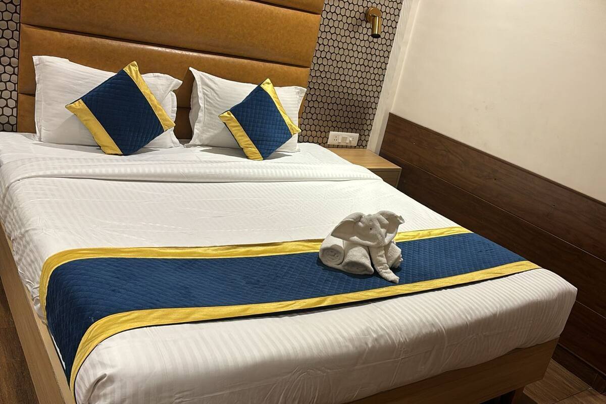 Habitación de hotel con una cama grande, dos almohadas, una colcha azul y amarilla, y una toalla enrollada sobre la cama.