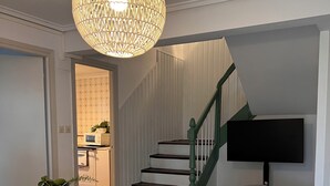 Un salon avec un canapé gris, un pouf rayé, une petite table d’appoint blanche ornée d’une plante, un grand luminaire tressé, un escalier avec des rampes vertes et un téléviseur à écran plat fixé au mur.