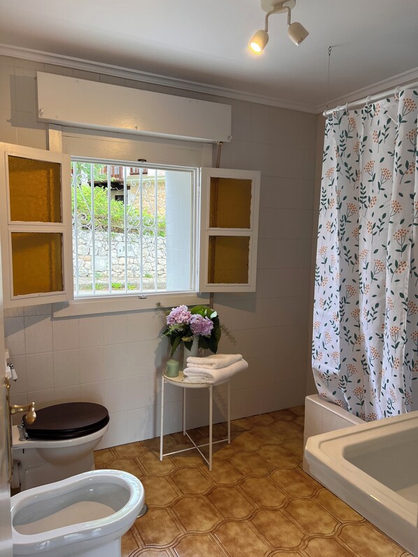 Un baño con inodoro, bidé, bañera, ventana con contraventanas decorativas y una pequeña mesa con toallas y flores.