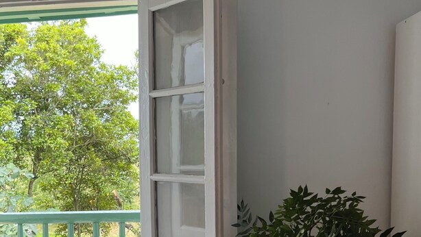 Une pièce donnant sur un paysage verdoyant, une porte blanche, une petite table blanche avec un vase de fleurs, un panier en osier contenant un coussin et une couverture posée sur le sol.