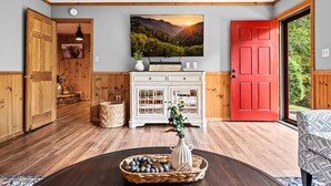 A spacious room with wooden floors, a red door, a white cabinet with glass doors, and a large window offering a view of greenery. - 3BR Gatlinburg*Firepit*Hot Tub*Privacy + Quiet! (Gatlinburg)