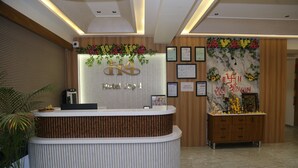 Un mostrador de recepción de hotel con diseño moderno, que incluye un logotipo, certificados enmarcados y una pared decorativa con arreglos florales.