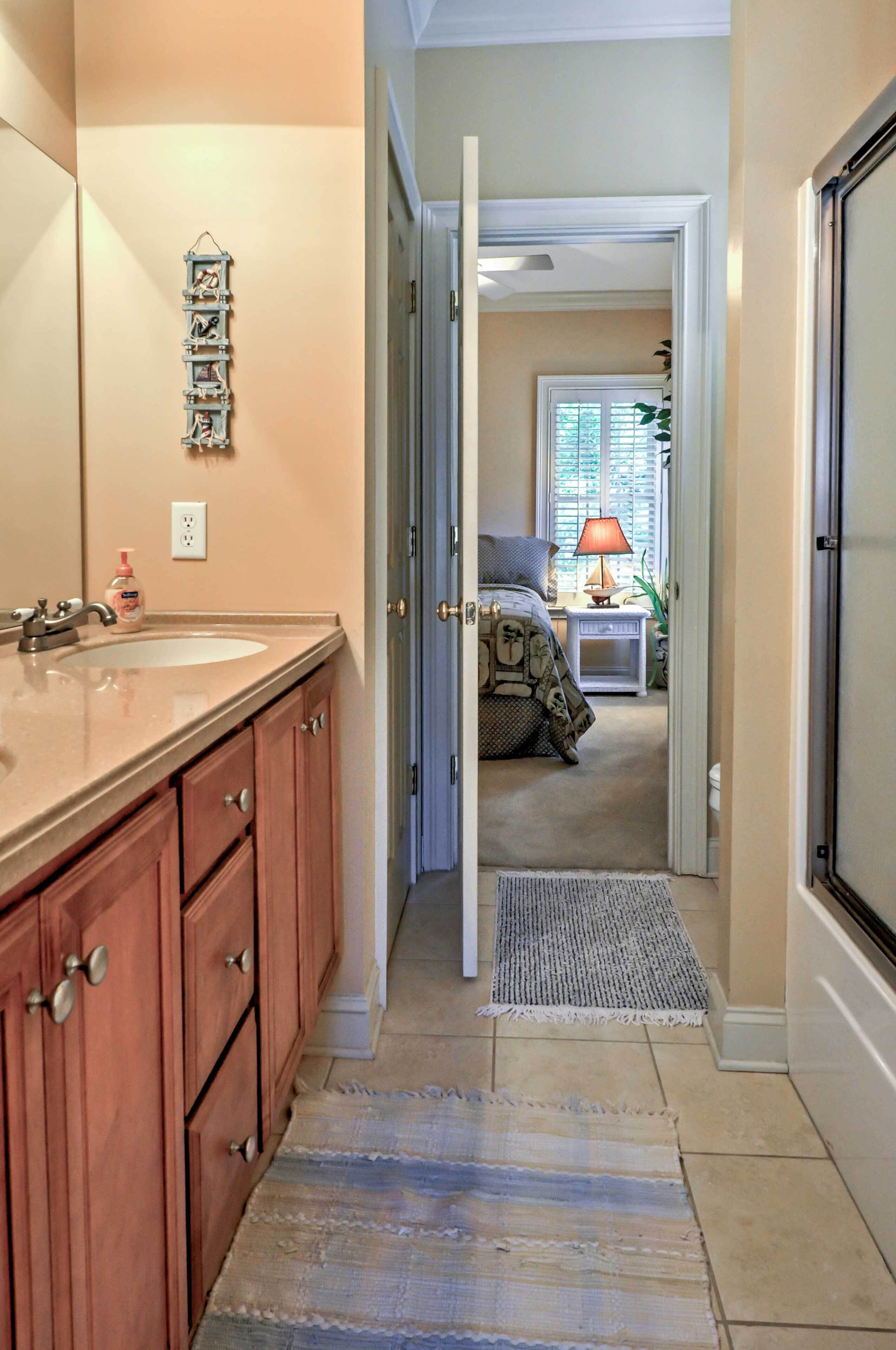 Une salle de bain équipée d’un lavabo, d’un miroir et de meubles en bois. Un couloir mène à une chambre comprenant un lit, une table de chevet et une lampe.