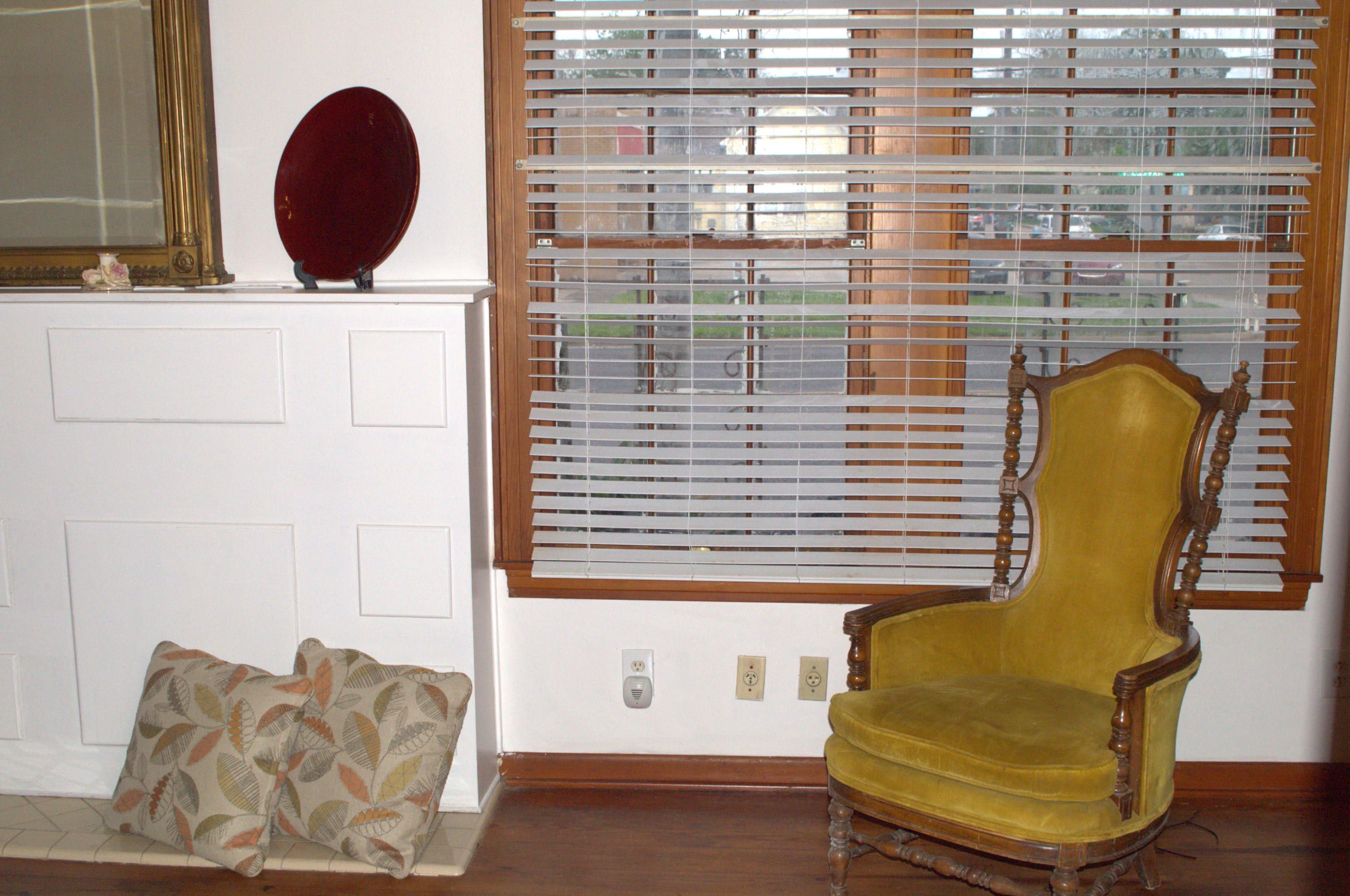 A room with a wooden-framed window with blinds, a yellow upholstered chair, a mirror, and a patterned cushion on a bench.