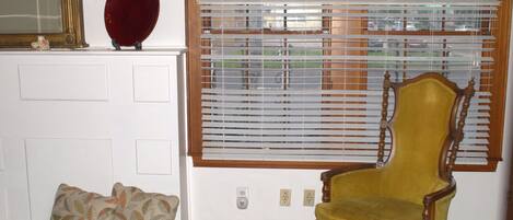 A room with a wooden-framed window with blinds, a yellow upholstered chair, a mirror, and a patterned cushion on a bench.
