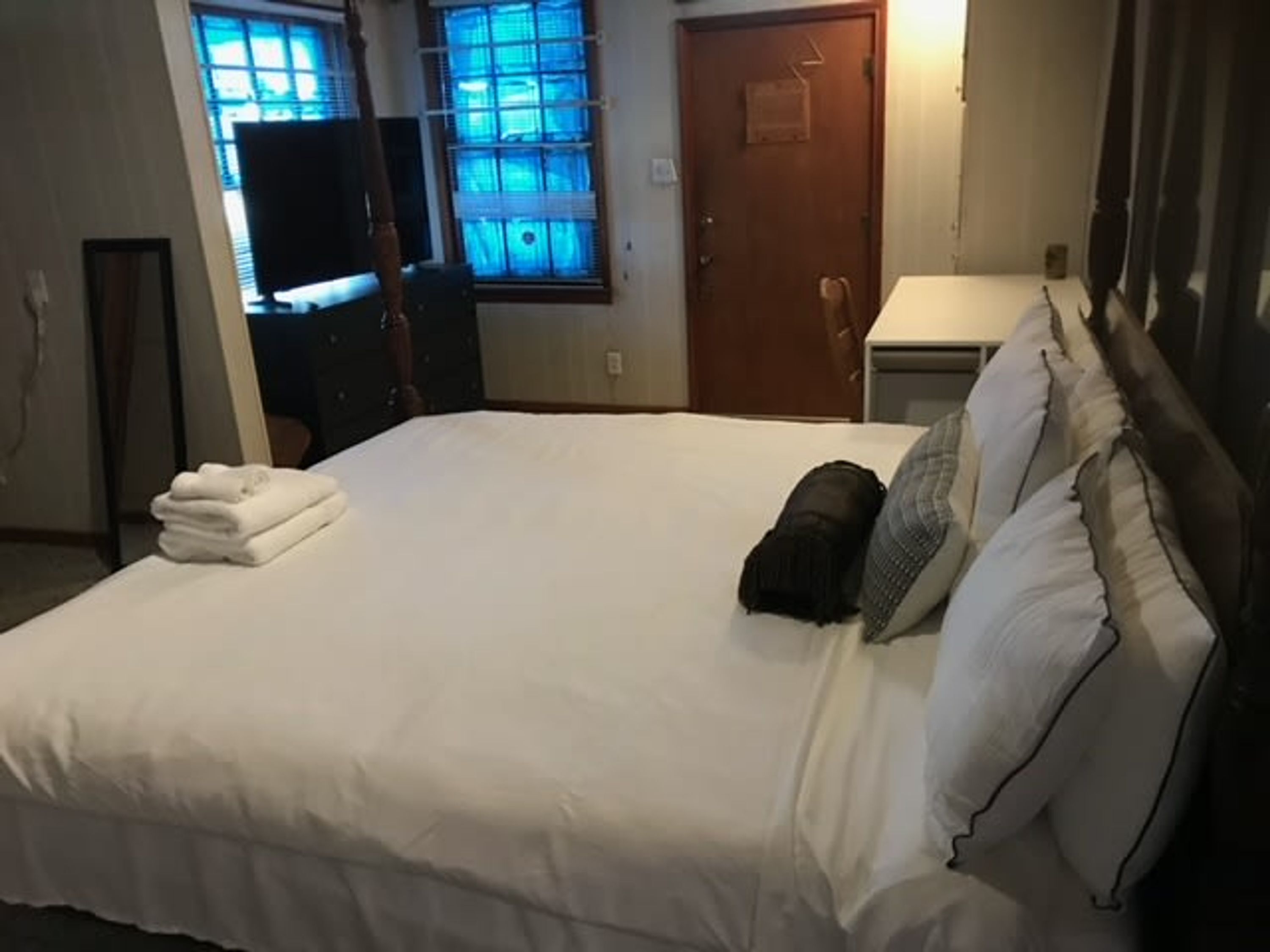A neatly made bed with white linens, a small black bag, and a pillow on a wooden headboard.