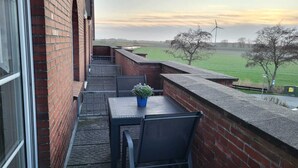 Un balcón con mesa y sillas, una planta en maceta y vistas a un campo verde y aerogeneradores.