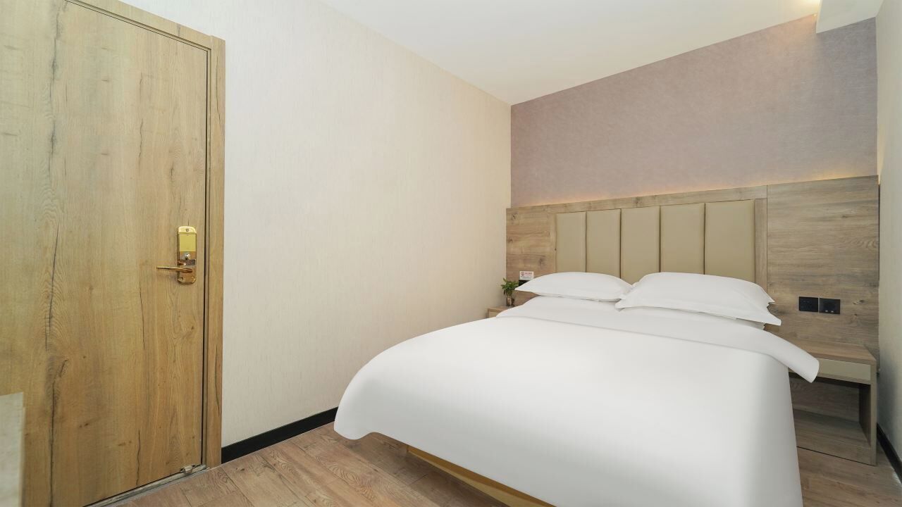 A hotel room with a wooden door, a bed with white linens, a headboard with built-in lighting, and a small bedside table.