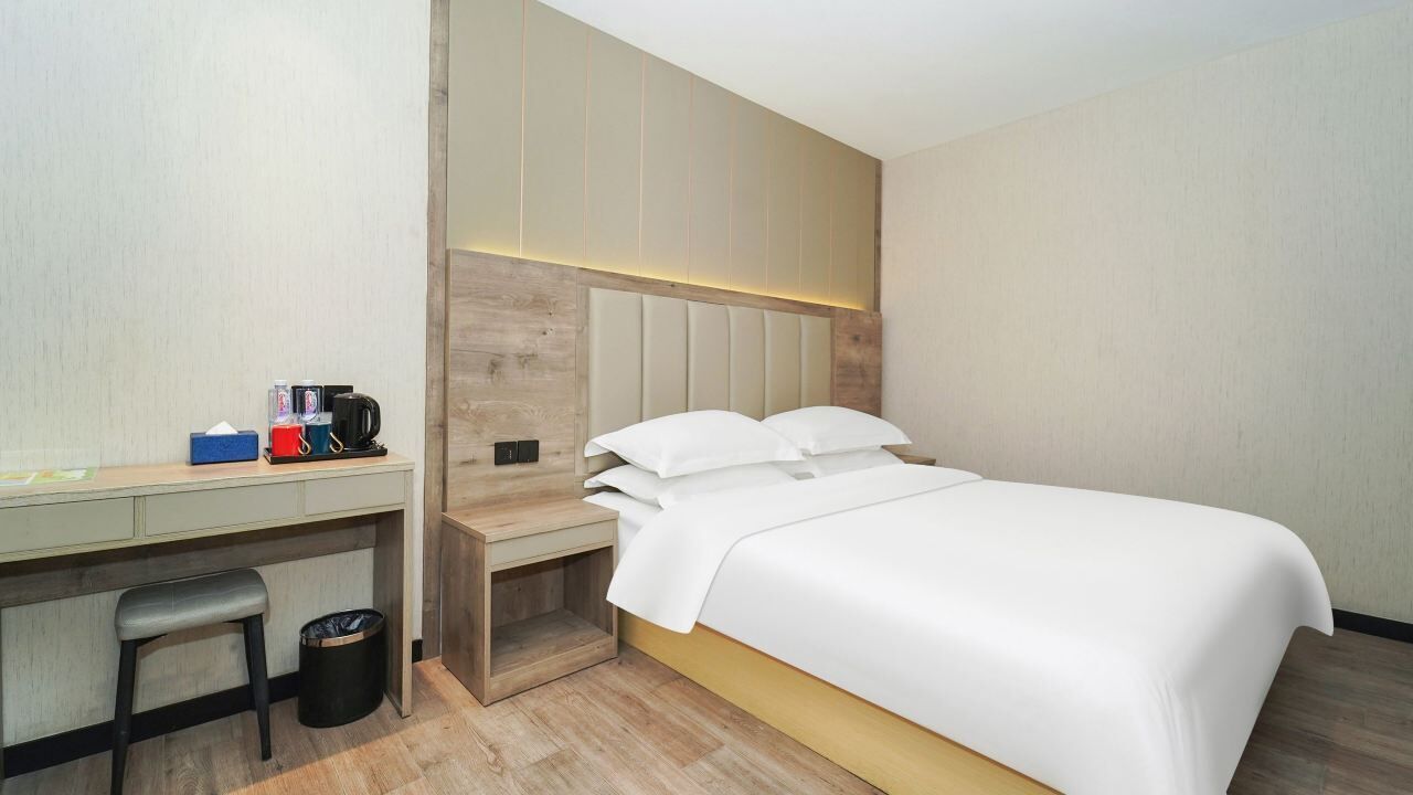 A modern hotel room with a bed, a desk, and a chair. There is a trash bin and a small table with a coffee maker and cups.