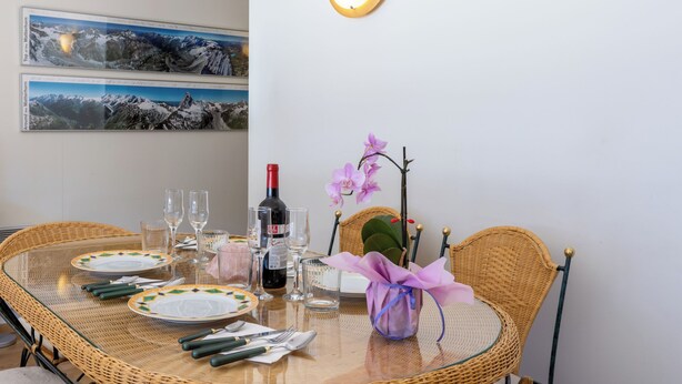 Une table à manger dressée avec des assiettes, des couverts, des verres et une bouteille, entourée de chaises en osier et d’un vase avec une fleur.