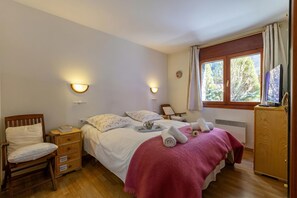 Um quarto com cama, mesas de cabeceira, uma cadeira, televisão e janela com cortinas.