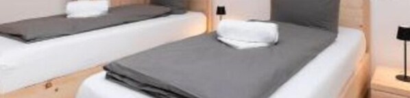 Ein Zimmer mit zwei Betten, jeweils mit Kissen und Decke. An einer Seite befindet sich ein Nachttisch mit einer Lampe.