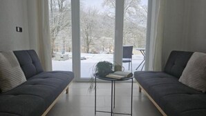 Ein modernes Wohnzimmer mit zwei dunklen Sofas, einem kleinen runden Tisch mit einem Buch und einer Pflanze, und Blick auf eine verschneite Landschaft durch große Fenster.