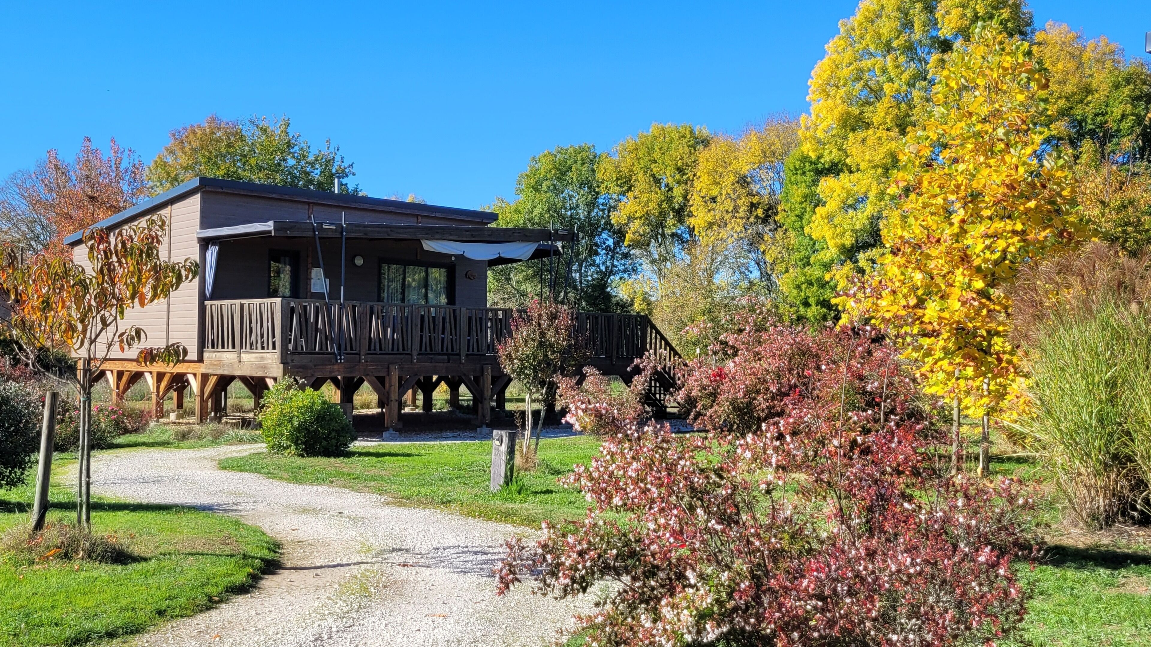 A wooden cabin with a deck, surrounded by a gravel path and various trees.