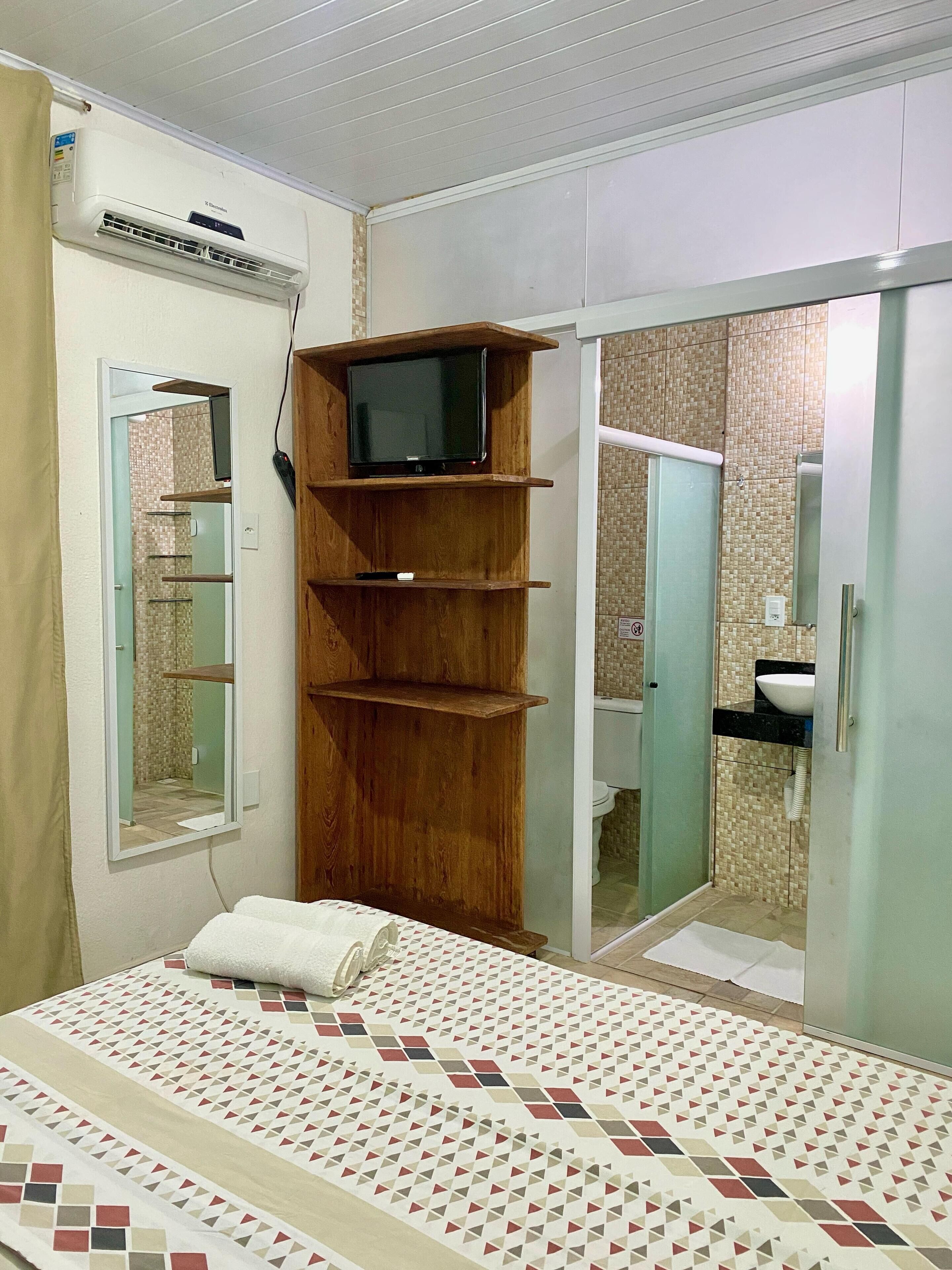 A bedroom with a bed, a wooden TV stand, a mirror, and a bathroom visible through an open door.