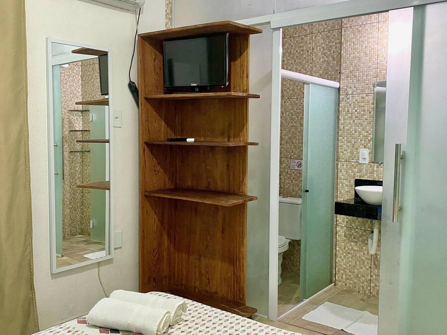 A bedroom with a bed, a wooden TV stand, a mirror, and a bathroom visible through an open door.