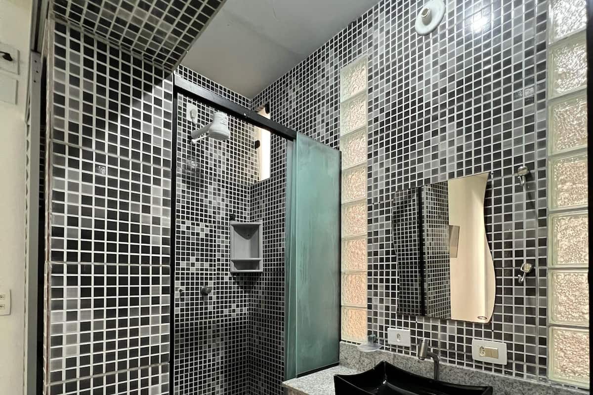 Un baño moderno con ducha acristalada, un lavabo con lavamanos negro y un espejo con lámpara sobre él.