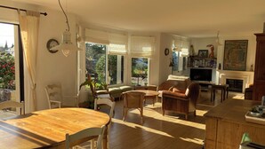 A spacious living room with a wooden dining table, comfortable seating, a fireplace, and large windows offering a view of the garden. - Spacious and bright house on large plot 10 min walk from Pont L' Abbé (Pont-l'Abbé)