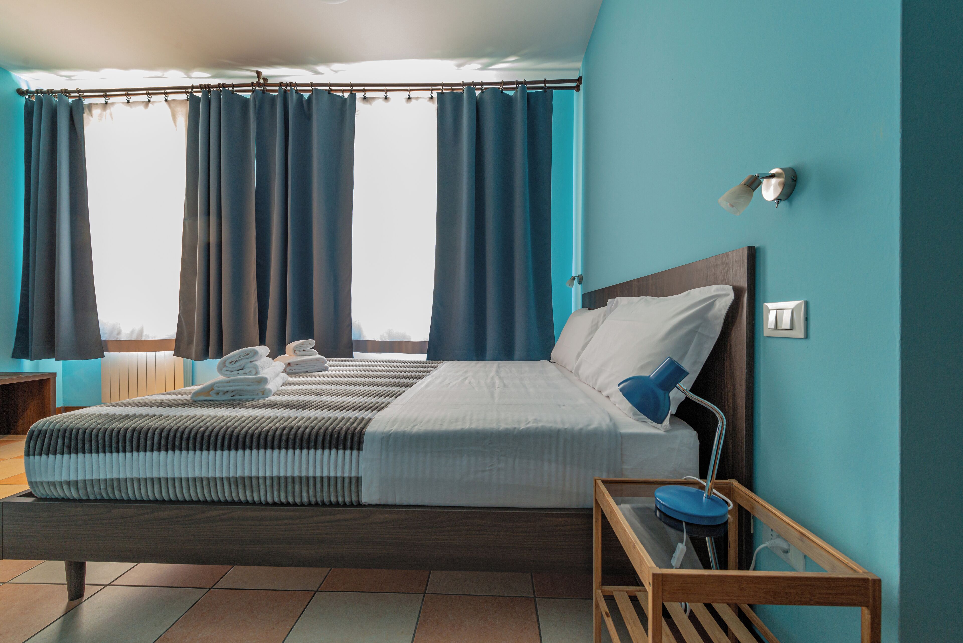 Un hotel moderno con una cama grande, una mesita de noche, una lámpara y vistas de la ciudad a través de amplios ventanales.
