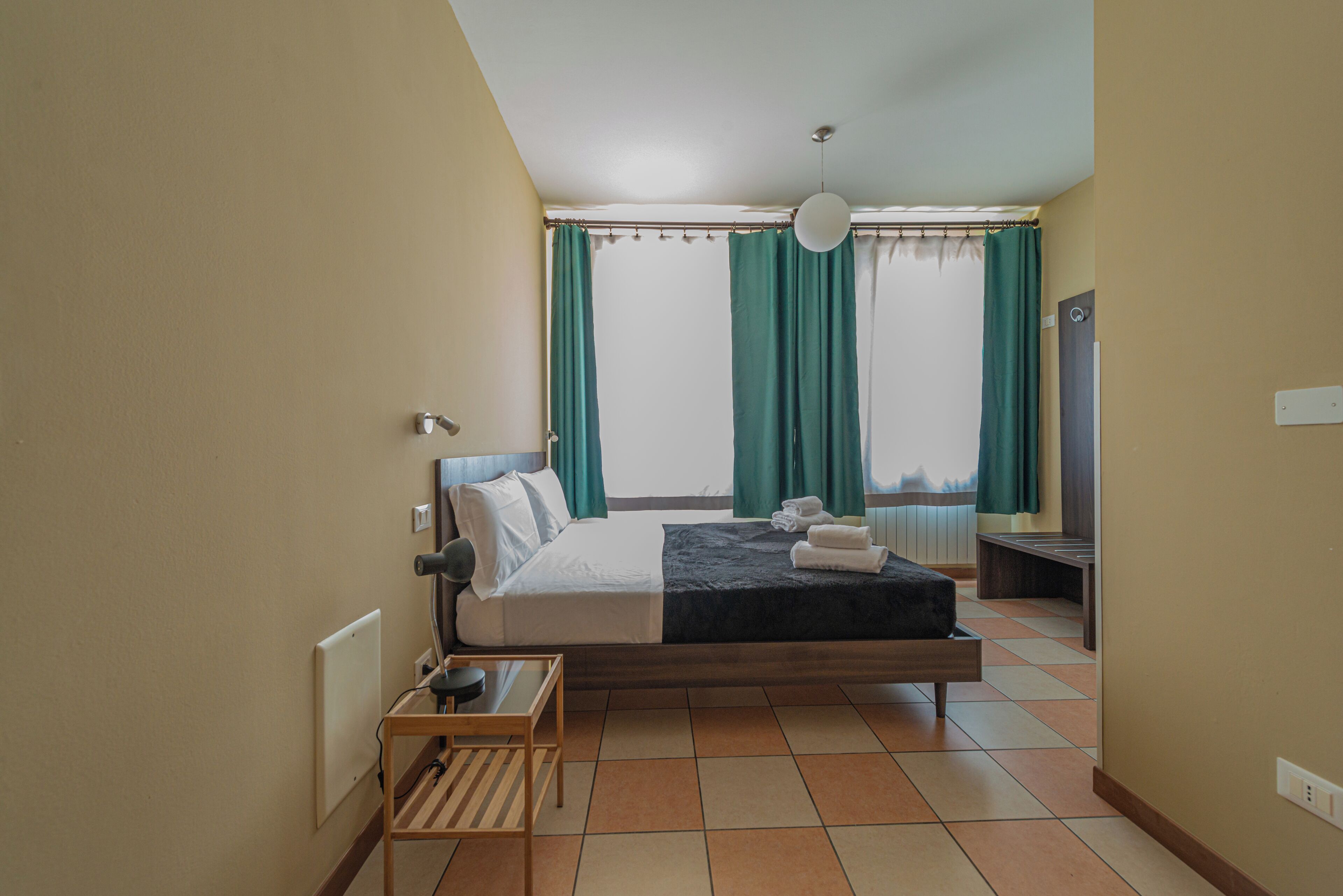 Habitación de hotel con cama, mesita de noche, silla y vistas a la ciudad a través de amplios ventanales.