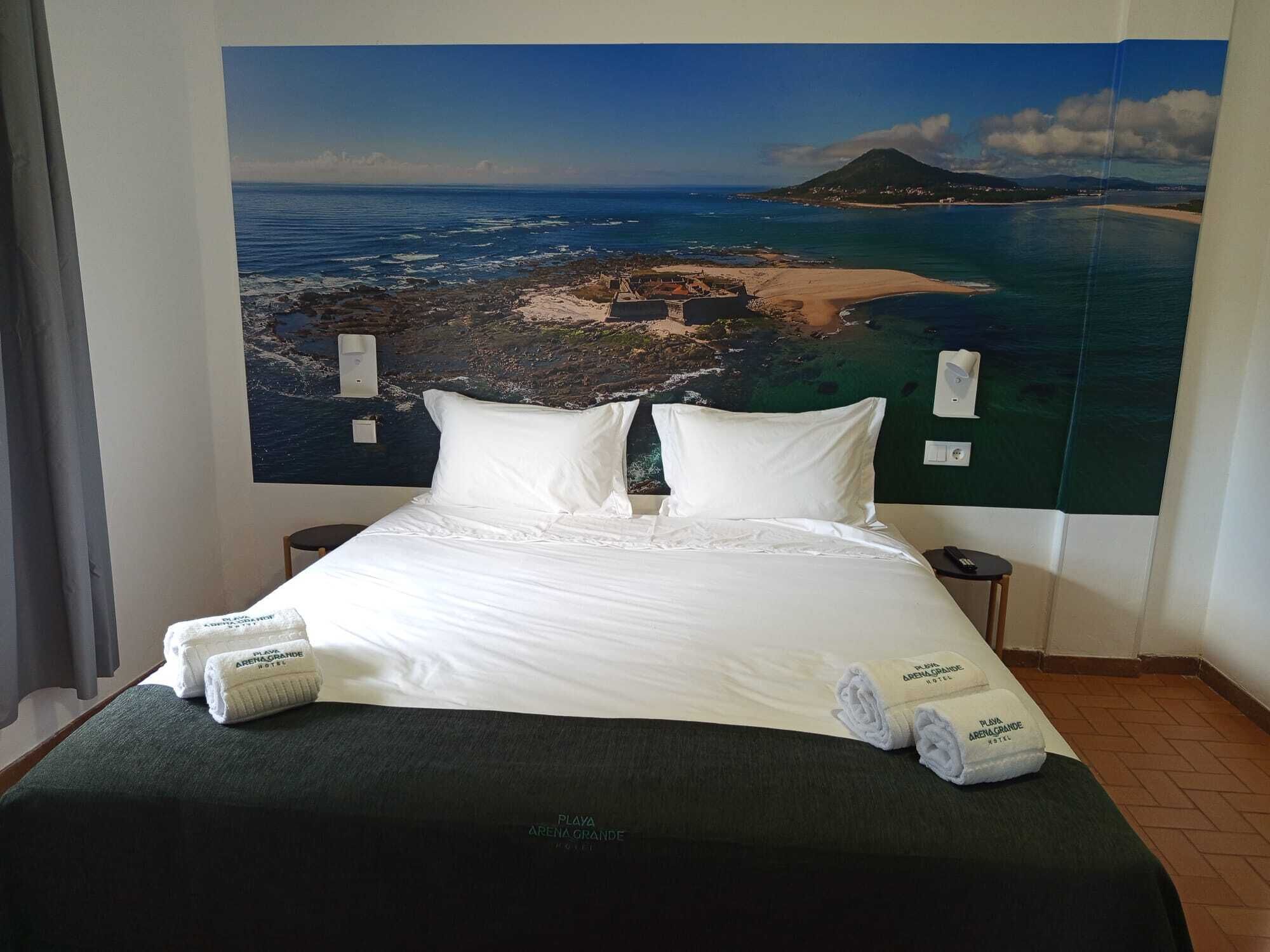 Ein Hotelzimmer mit einem Bett, zwei Kissen, einem Nachttisch, einer Lampe und einem großen Wandgemälde einer Küstenlandschaft.