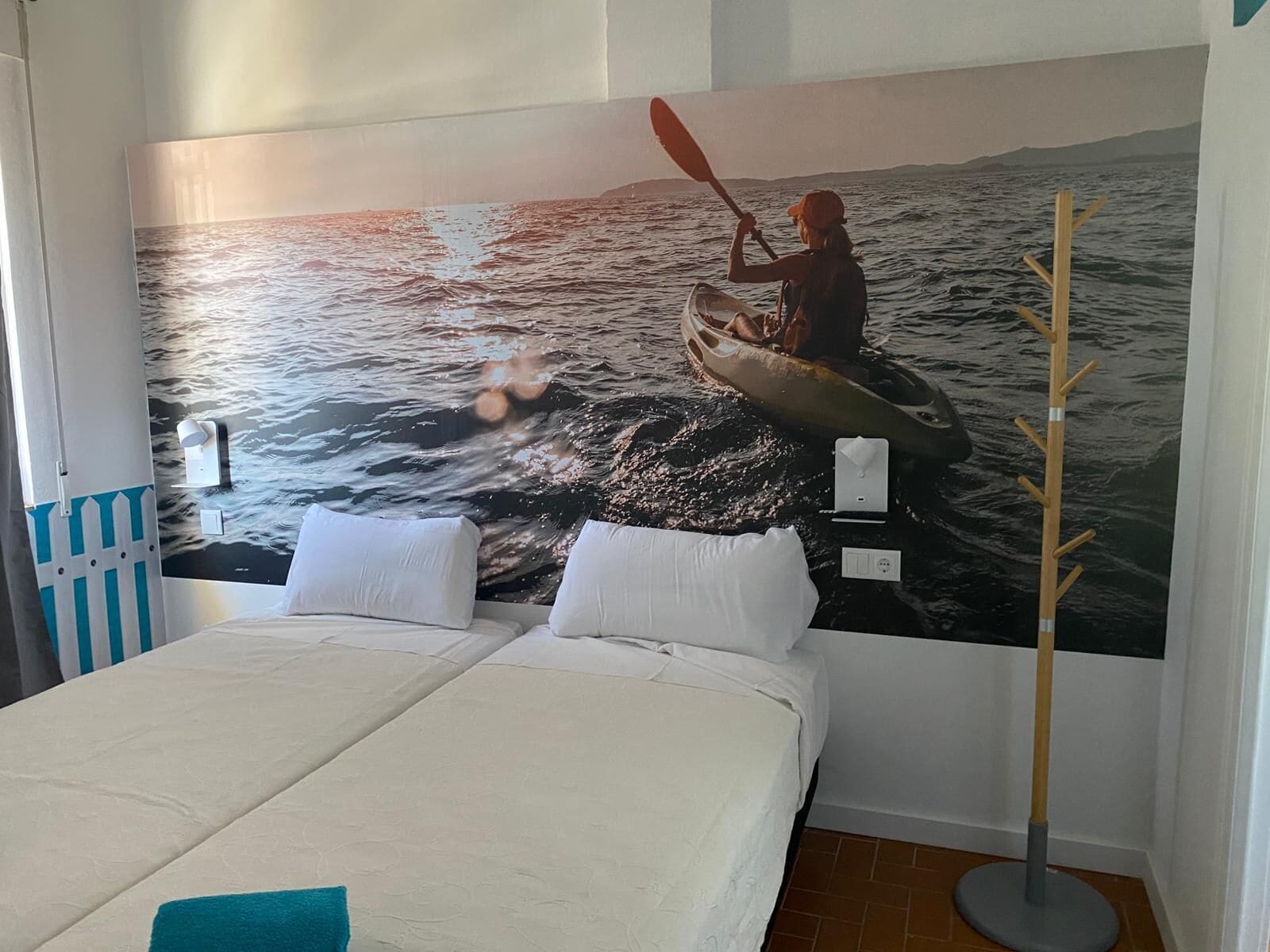 A bedroom with a large mural of a kayaker on water, a bed with white linens, and a wooden coat rack.