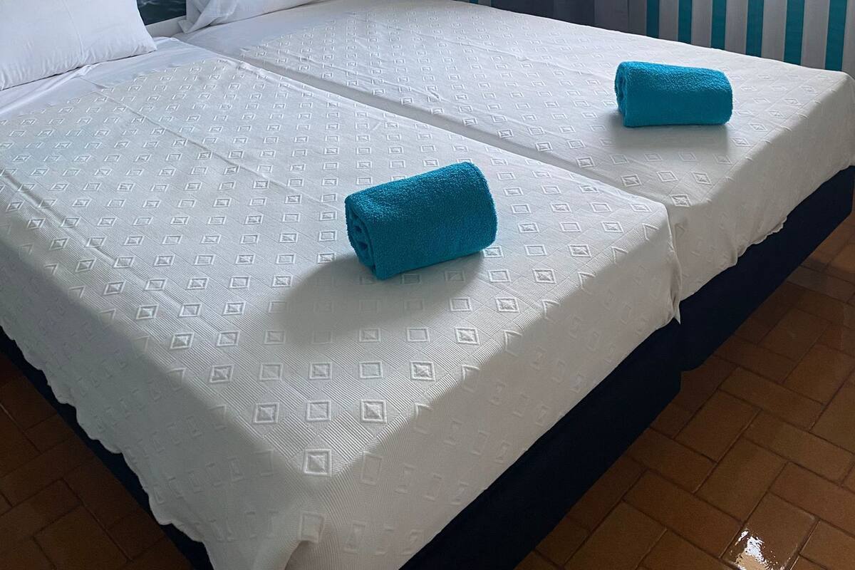 A hotel room with a large bed, two pillows, a blue towel, a wall mural of the ocean, a window with curtains, and a white picket fence design on the wall.