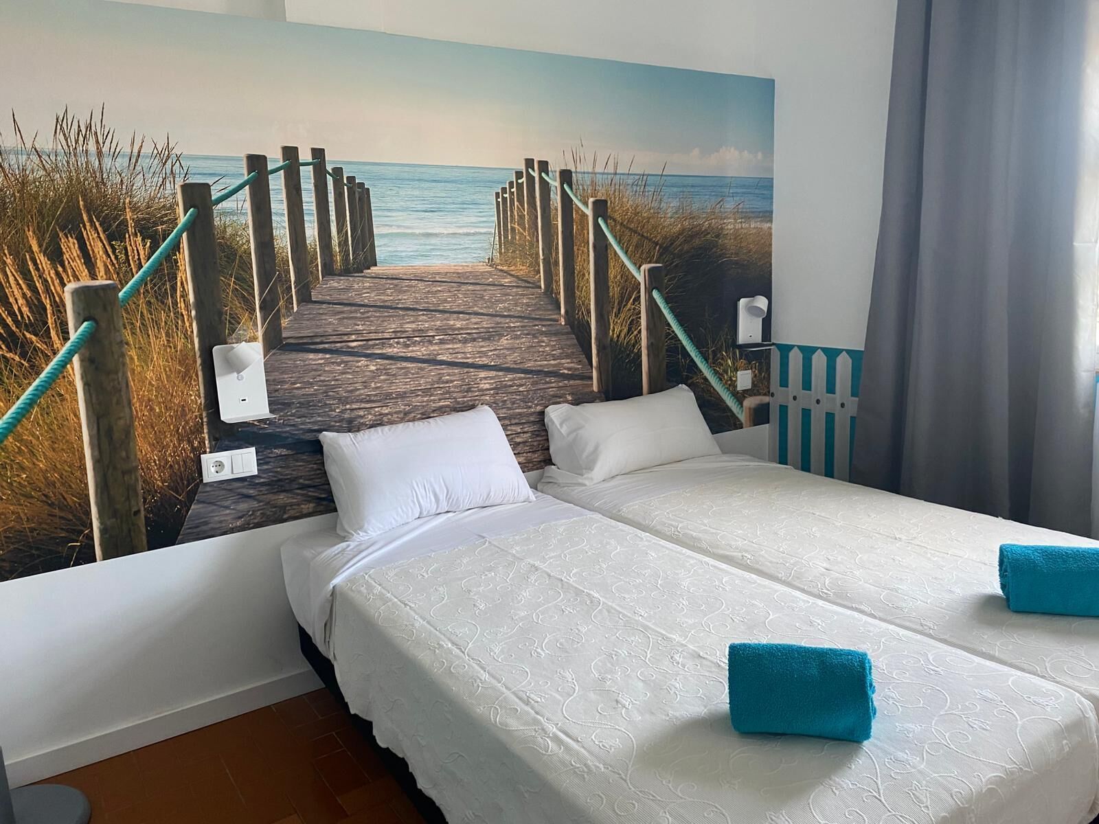 A hotel room with a bed, two pillows, a blue towel, and a mural of a pier leading to the sea.