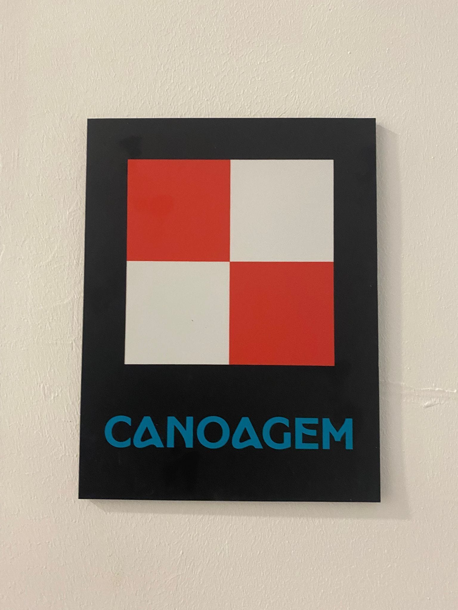 Un letrero con un diseño cuadrado rojo y blanco y la palabra "CANOAGEM" en letras azules.