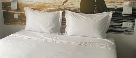 A bed with white linens and pillows, a wall mural of a woman at the beach, and two towels with "Playa Arena Grande Hotel" text.