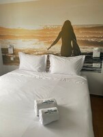 Una cama con sábanas y almohadas blancas, un mural en la pared de una mujer en la playa, y dos toallas con la inscripción "Playa Arena Grande Hotel".
