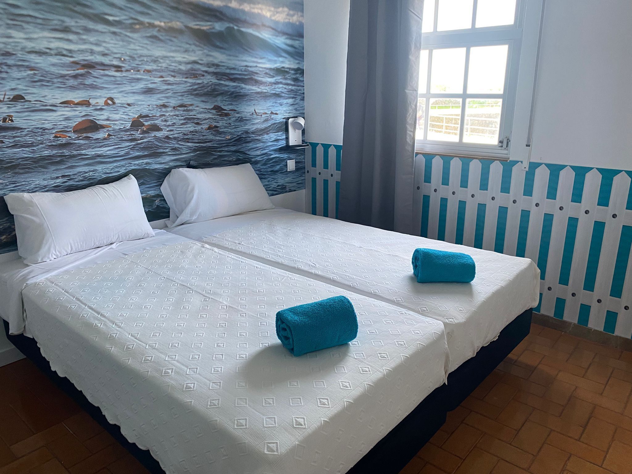 A double bed with white linens and two blue towels, a mural of the ocean on the wall, a window with curtains, and a white picket fence.